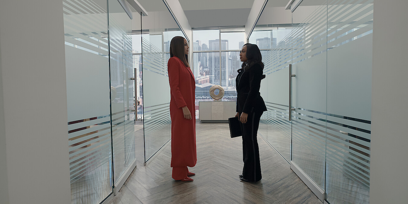 Crystle Stewart as Mallory and Taylor Polidore Williams as Kimmie in business attire stand facing each other in a modern office hallway with glass walls, city skyline visible through large windows in the background.