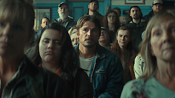 A group of people sit closely together indoors, appearing attentive and serious, likely at a community meeting in a rustic room with blue walls and casual clothing, with the central man in focus.