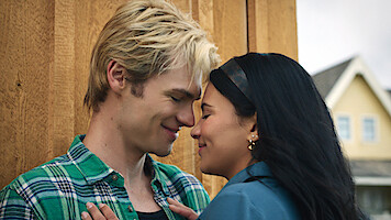 A young couple stands close together, smiling with their foreheads touching in an intimate moment outside near a wooden wall and houses in the background.