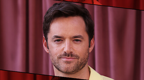 A man with short dark hair and a trimmed beard is standing in front of pink curtains. The setting appears indoors with soft lighting, and the image is framed with red and black graphic borders.