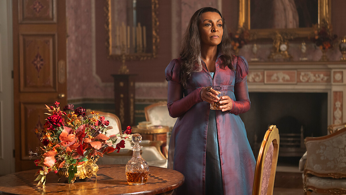 Woman in a purple Regency dress stands by a table with flowers and a decanter in an ornate, classic room with a fireplace, mirror, and rich decor, holding a glass and looking thoughtful.