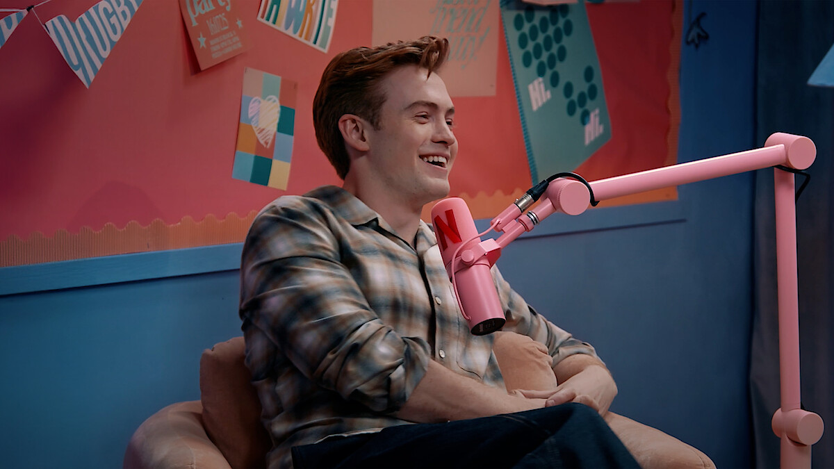 Kit Connor sitting and talking into a pink microphone in a colorful studio with artwork and notes on the wall behind them.