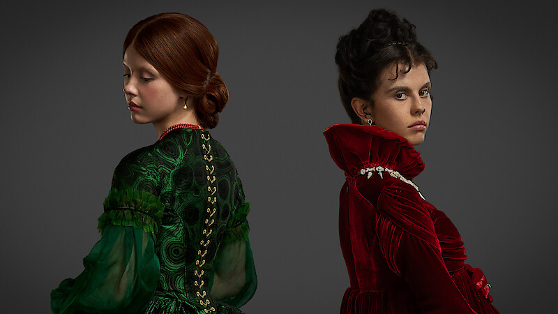 Two women in elaborate period costumes, one in green and one in red, stand back to back against a dark background. Their serious expressions and elegant dresses create a dramatic, tense, and formal mood.