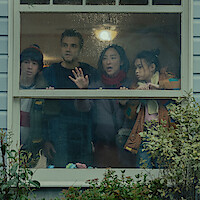 Four people stand closely together indoors, looking anxiously out a rainy window of a house with pale siding, surrounded by green shrubs, under soft indoor lighting.