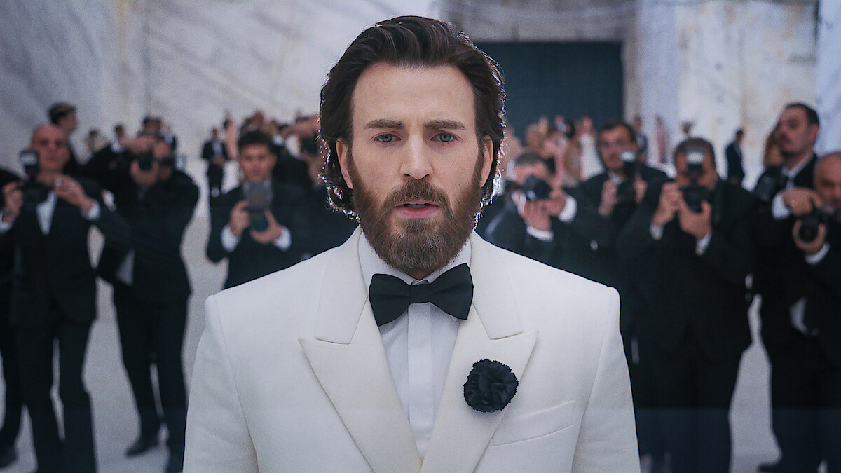 A bearded man in a white tuxedo stands in a marble room, surrounded by photographers in black suits taking his picture, creating a formal and glamorous atmosphere.