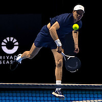 Tennis player in navy outfit serving on indoor hard court, with a yellow tennis ball in mid-air and a Riyadh Season logo visible in the background.