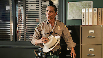 Smiling sheriff in uniform holding white hat stands in an office with filing cabinets, folders, a wall map, and windows with blinds in the background.