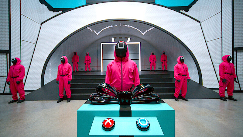 Guards in pink uniforms and masks stand in a futuristic, sterile room; one stands behind a control panel with red and blue buttons, creating a tense, mysterious atmosphere.