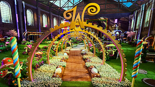 A whimsical candy-themed garden inside a large factory with colorful sweets, oversized mushrooms, lollipops, golden arches, flowers, and a large "W" centerpiece under a glass roof, creating a magical, playful atmosphere.