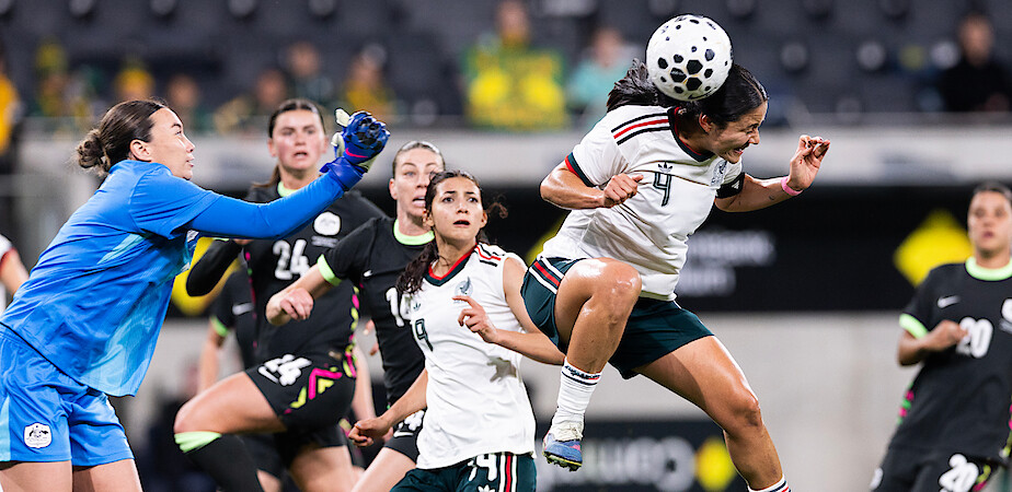 Women's soccer match in a stadium, goalkeeper in blue reaches for the ball as player in white and green jumps to head it, teammates and opponents in action, focused expressions, blurred spectators in background.