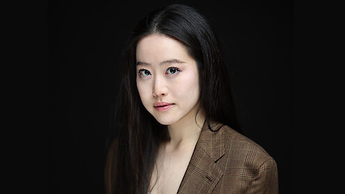 Woman with long black hair in a brown checked blazer poses against a plain black background, softly lit, looking confidently at the camera in a professional studio portrait setting.