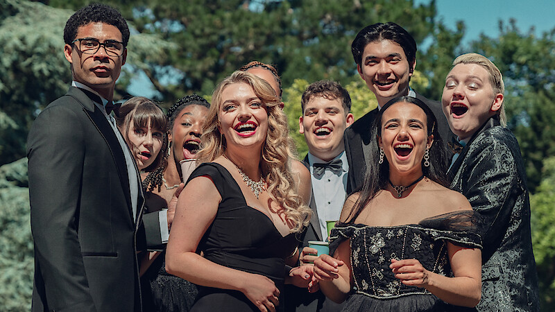 A group of people in formal attire stand outdoors, smiling and laughing together in a lush, green, sunlit setting, suggesting a celebratory or special event.