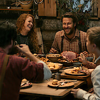 Four people share a rustic meal in a cozy, wooden cabin. The table is set with hearty food, and everyone is smiling and laughing, suggesting a warm, joyful gathering in a homey, inviting environment.