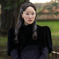 A woman in Victorian-style black clothing sits outdoors in a park or garden, looking concerned while talking to another person dressed similarly. Trees and a fence are visible in the background.