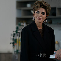 Woman with short styled hair and hoop earrings wearing a belted black blazer stands indoors in a modern room, medical equipment blurred in the background, while someone hands her a white card or envelope.