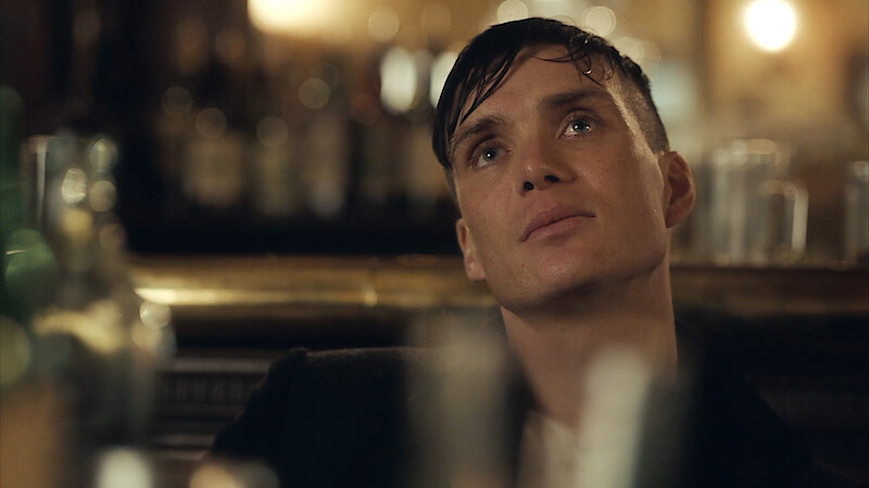 Man with slicked hair sits in a dimly lit bar, gazing upward thoughtfully. Glasses and bottles are blurred in the foreground; soft, warm lighting and an out-of-focus background create a moody atmosphere.