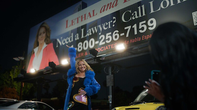A woman in a blue coat poses under a large lawyer advertisement billboard at night while another person takes her photo. Cars and city lights are visible in the background, creating an urban nighttime scene.