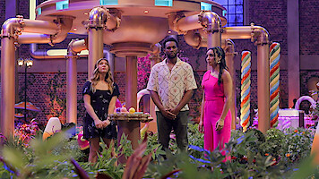 Three people stand in a whimsical, candy-themed indoor set with bright decor, oversized pipes, and colorful candy cane props, surrounded by plants and soft lighting, giving a playful, fantasy atmosphere.