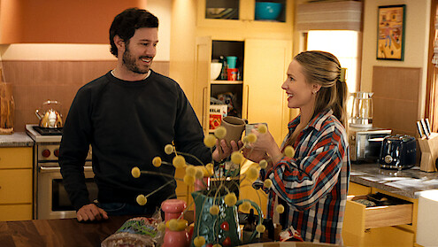 Two people smiling and toasting mugs in a cozy, brightly lit kitchen with yellow cabinets, assorted groceries on the counter, and decorative items in a homey setting.