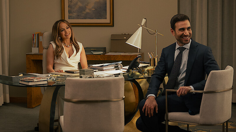 A woman and a man in business attire sit and smile in a modern office with a glass desk, papers, books, and a desk lamp, with neutral-colored walls and a framed picture in the background.