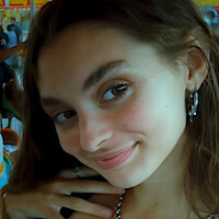 Young woman smiling for a selfie on a merry-go-round with carousel horse and people in the background, indoor amusement setting, colorful and lively atmosphere.