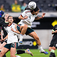 Female soccer player in white jersey jumps and heads the ball during a match; opponents and teammates are nearby on a professional field with blurred stadium background.