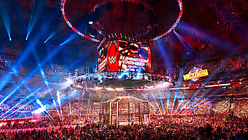 WWE Elimination Chamber wrestling event in a large arena, filled with a cheering crowd, colorful lights, a cage structure in the ring, and giant display screens with event logos and a WrestleMania sign in the background.