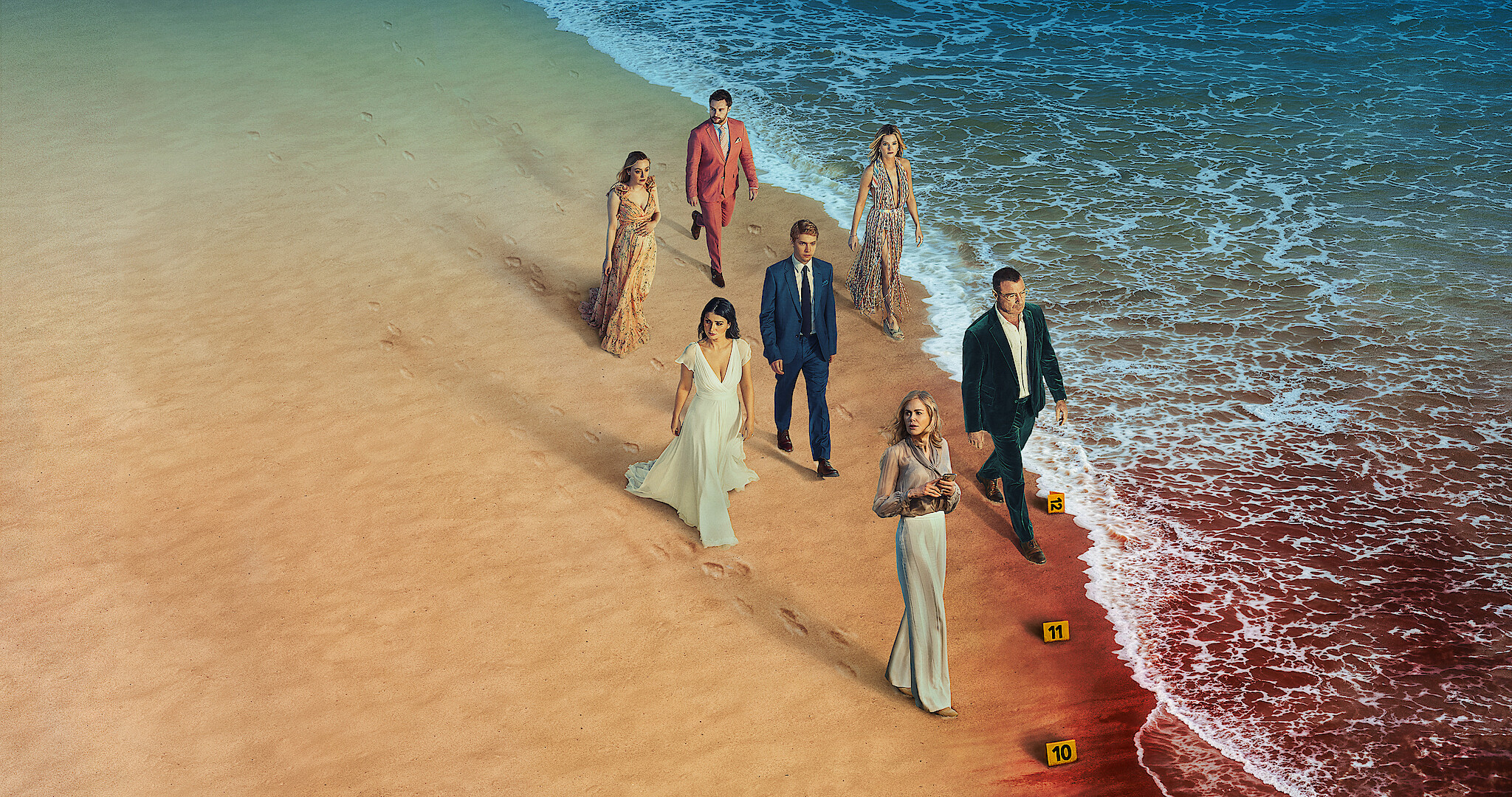 The cast of the Perfect Couple walk along a beach next to the ocean, with a section of it tinged red with what looks like blood. 