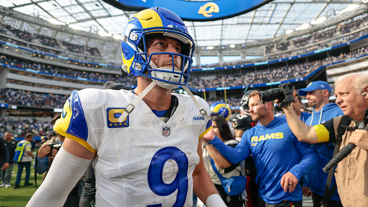 A Los Angeles Rams football player in uniform walks on the field at a stadium, surrounded by fans, photographers, and media, with the stadium roof and large crowd visible in the background.