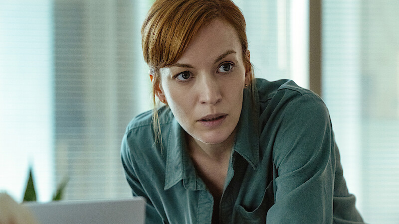 A woman with red hair in a green shirt leans forward in a modern office environment, looking intently at something off camera, with blurred windows and blinds in the background.