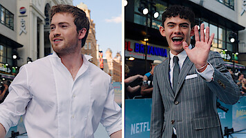 Kit Connor and Joe Locke at the Heartstopper Forever premiere, one in a white shirt smiling, the other in a pinstripe suit waving, with city buildings, lights, and Netflix banners in the background.