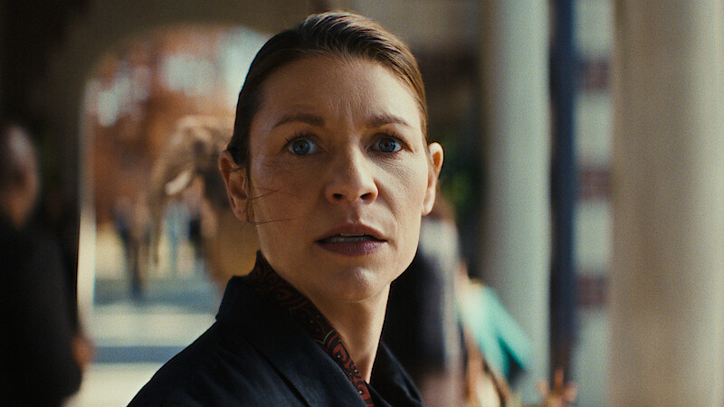 A woman with a serious expression stands outdoors in a sunlit area, with blurred people and architectural columns in the background, suggesting a public or urban setting.