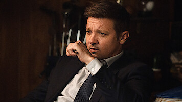 A man in a suit sits indoors, appearing deep in thought with his hand near his mouth, in a dimly lit room with wooden walls and blurred objects in the background.