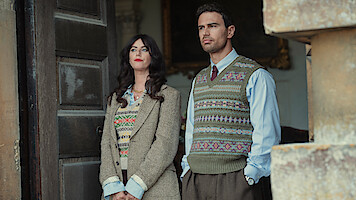 A woman and a man in vintage sweater vests and formal clothing stand side by side in front of a large open door, with stone architecture visible, looking thoughtfully into the distance.