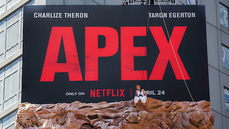 Large outdoor billboard for "APEX" on Netflix with bold red text, release date, actor names, and a person in a harness suit sitting on a rock-like structure in an urban cityscape.