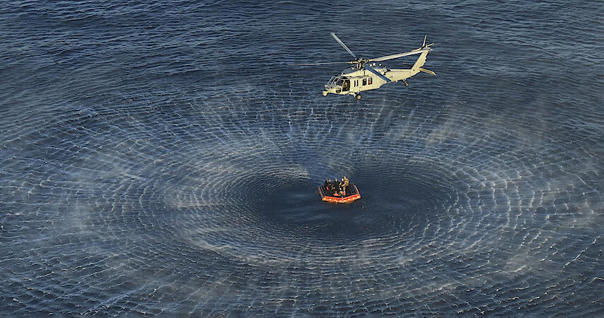 A helicopter hovers above the ocean, creating circular waves while performing a rescue operation near a life raft with several people aboard.