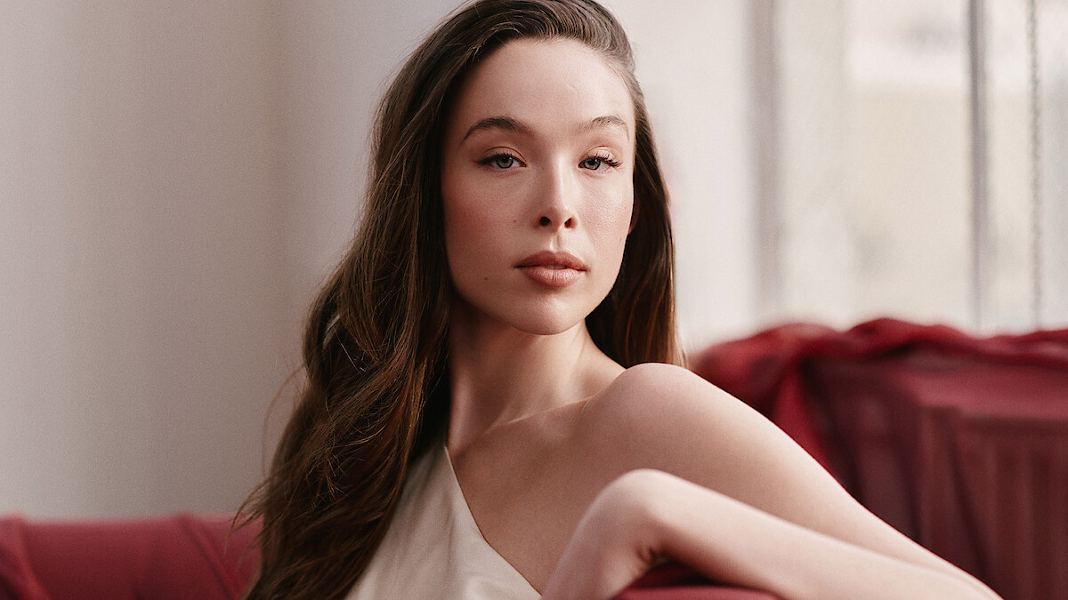 Ella Beatty wears a white dress while sitting on a chair draped with red fabric.