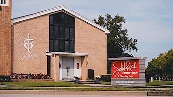 Brick church building with large black window, cross and heart logo, and "Solid Rock" sign out front. Well-maintained lawn, flowers, and trees in a suburban environment under a clear sky.