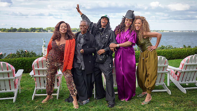 Five stylish women stand outdoors on green grass in front of white chairs by a lake, smiling and posing happily under a partly cloudy sky.