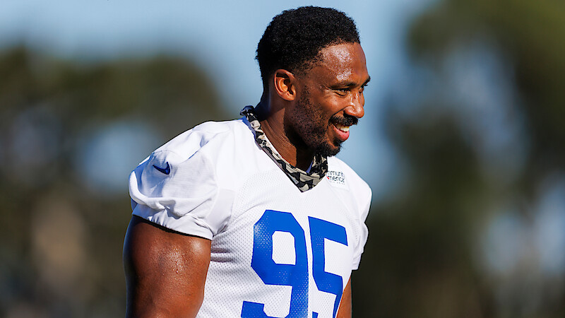 Football player wearing a white jersey with the number 95 smiles during an outdoor practice session, with blurred trees and blue sky in the background.
