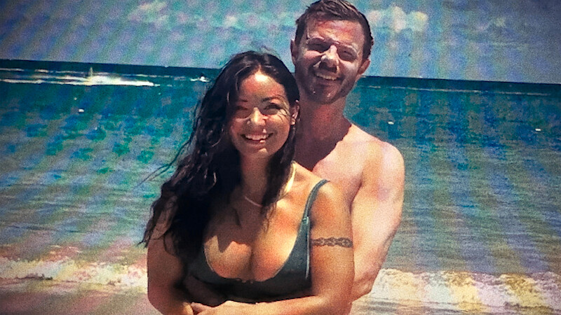 A smiling couple standing together on a sunny beach with waves and a blue ocean in the background.