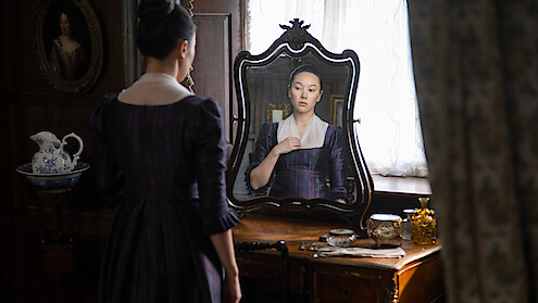 A woman in historical clothing stands at a wooden vanity, looking at her reflection in an ornate mirror, with antique items and soft natural light filling the elegant, vintage room.