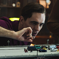 A man in a library or study leans over a detailed model of a bridge with miniature cars and people, carefully arranging the scene with his hand.
