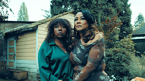Two women stand close together outside beside a rustic shed, surrounded by greenery and trees. Both look thoughtful, dressed formally, with overcast natural lighting creating a serene, introspective mood.