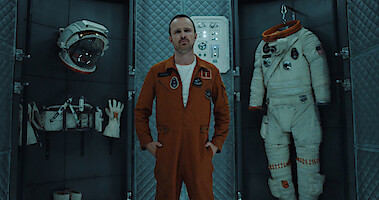 Aaron Paul as XX wears an orange jumpsuit with an American Flag patch while sitting in front of a control panel in Season 6 of Black Mirror.
