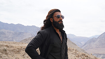 Man with long hair and beard wearing sunglasses and dark clothing stands confidently in a rugged mountainous landscape under a cloudy sky, suggesting a cool and stylish vibe in a remote outdoor environment.