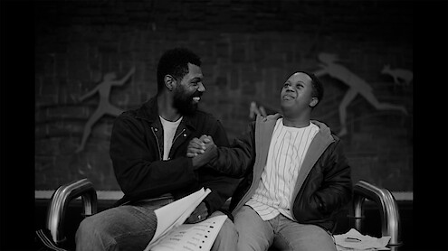 Two men sitting on a bench indoors, smiling and clasping hands in a friendly gesture, with papers on their laps and athletic figures depicted on the wall behind them.