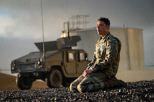 A soldier in camouflage uniform kneels on rocky ground near a military vehicle, with an industrial tank in the background under a cloudy sky, capturing a contemplative and solemn mood.