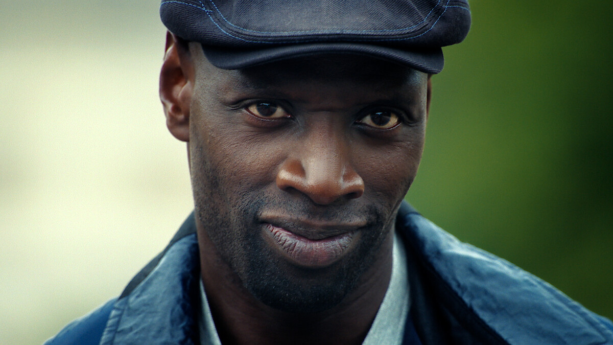 A man outdoors wearing a blue jacket, light shirt, and dark flat cap, looking directly at the camera with a subtle, confident smile and a blurred green background.