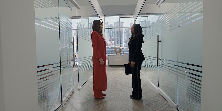 Crystle Stewart as Mallory and Taylor Polidore Williams as Kimmie in business attire stand facing each other in a modern office hallway with glass walls, city skyline visible through large windows in the background.